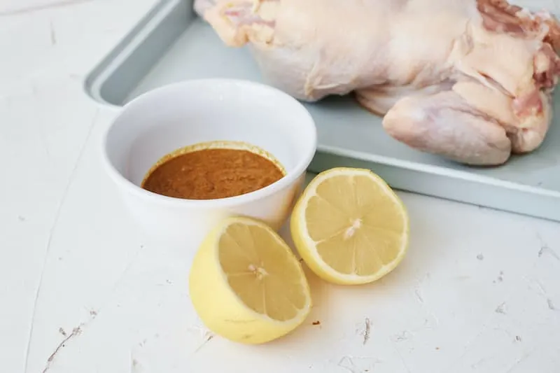 Whole raw chicken on a tray with a lemon cut in half and spice mixture in the foreground.