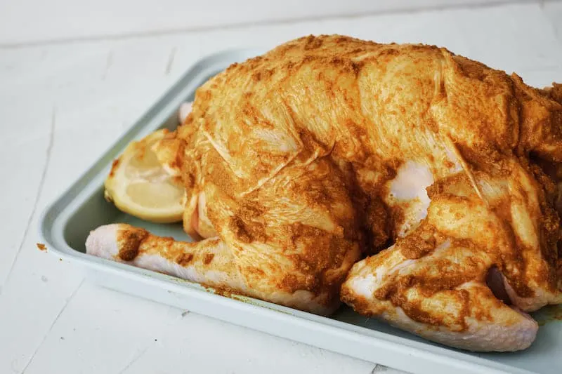 Whole raw chicken coated in seasoning marinade on a baking tray.