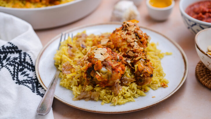 A white plate of yellow rice is topped with harissa baked chicken.