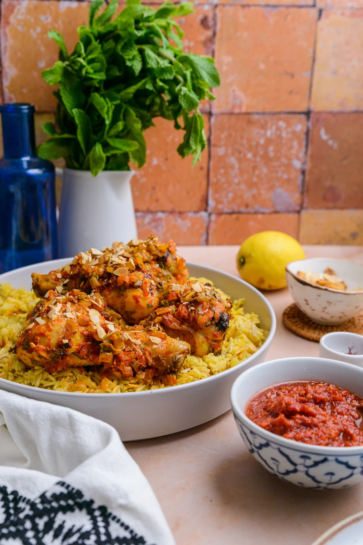 Several bowls sit on a table, the large bowl in the background has saffron rice and is topped with pieces of chicken coated in harissa sauce.