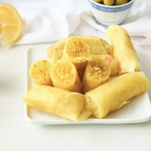 Stacked Moroccan briouats wrapped in a cigar shape with lemons and dipping sauce in the background
