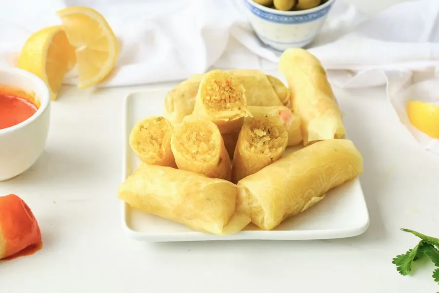 Stacked Moroccan briouats wrapped in a cigar shape with lemons and dipping sauce in the background