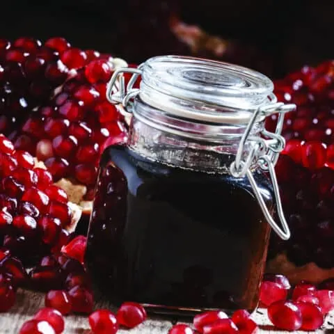 Glass bottle of pomegranate syrup with a flip top lid in the front of the image and cut open pomegranates in the background.