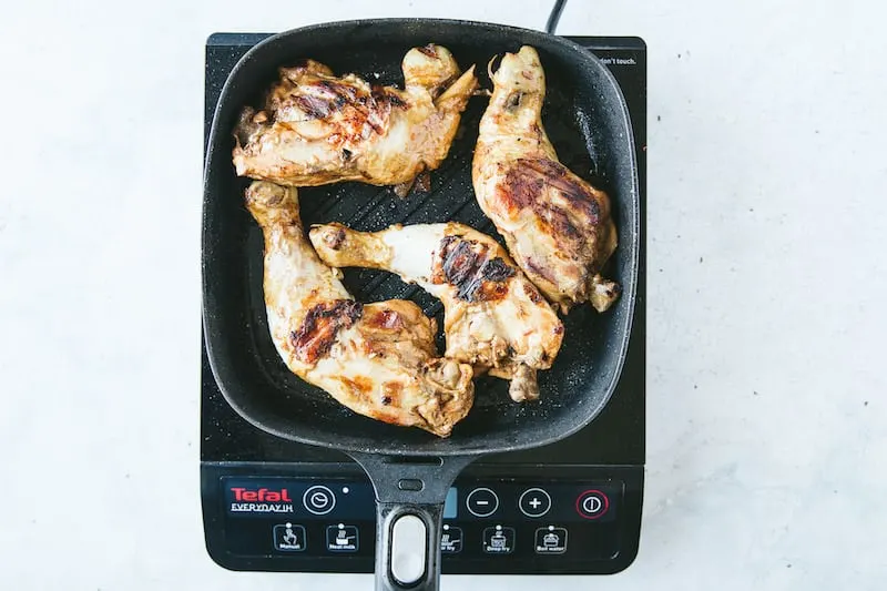 A grill pan is on a stovetop with chicken pieces cooking