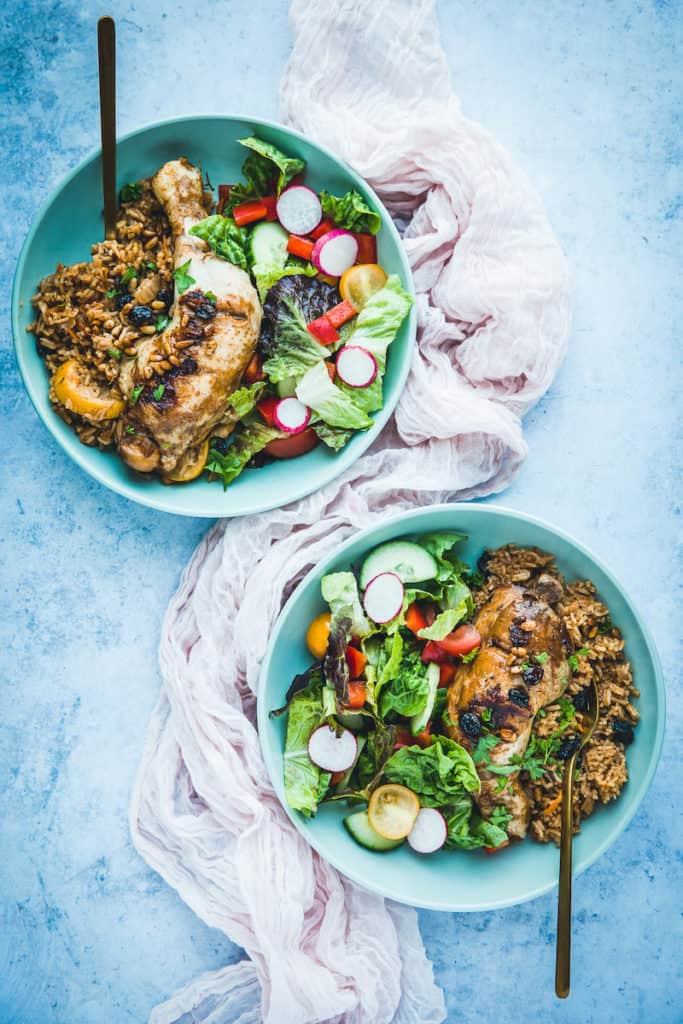 Two bowls of chicken kabsa on a blue background. There is salad and chicken in the bowl.