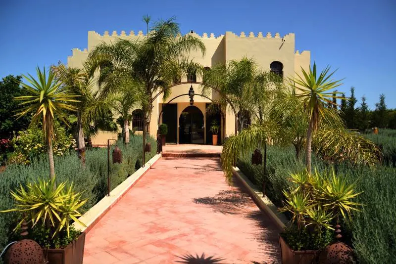 A red pathway leads to a sand colored castle style building in Fez. Green trees and bushes surround the path and building.