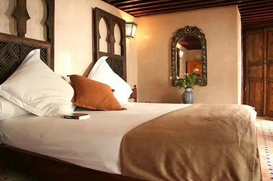A large bed is covered with white linen and a brown blanket. The room of Riad Dar Dmana is large and cozy.