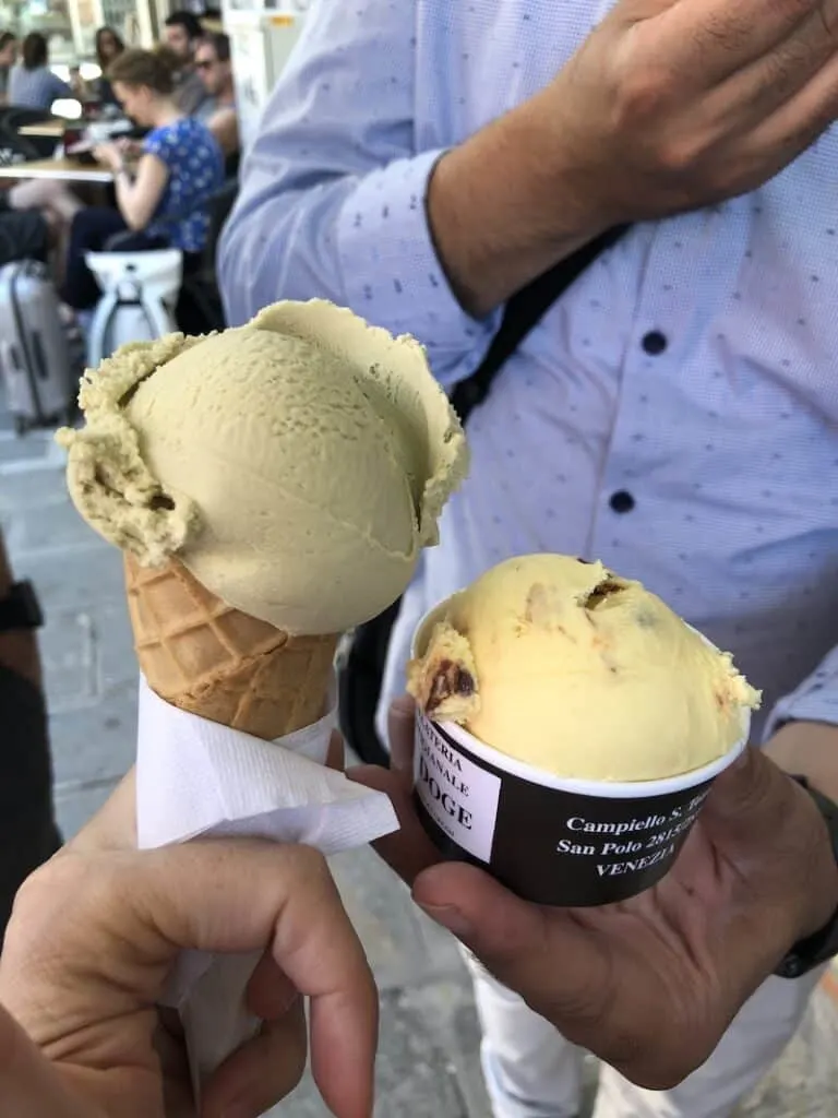 A man\'s hand holding a cup of nougat gelato. A woman\'s hand holding a cone of pistachio gelato.