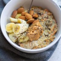 White bowl of carrot cake flavored oatmeal topped with bananas, almonds and peanut butter