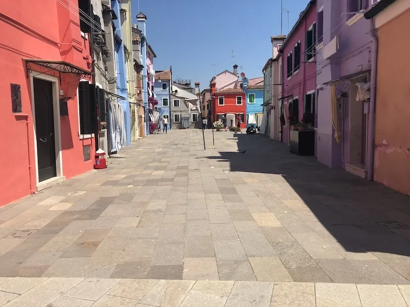 A pedestrian street in Burano Italy with brightly colored buildings on either side in violet, blues, and pinks.