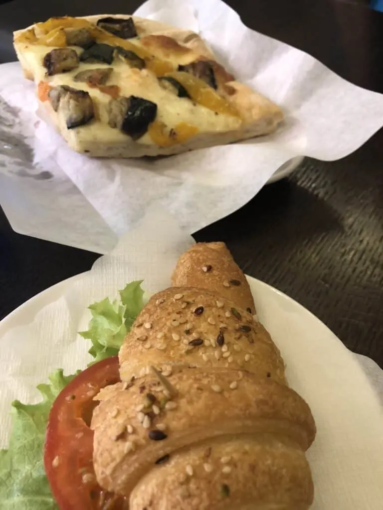A gluten free croissant with lettuce and tomato is in the front of the picture and a slice of pizza with cheese and eggplants sits behind it.