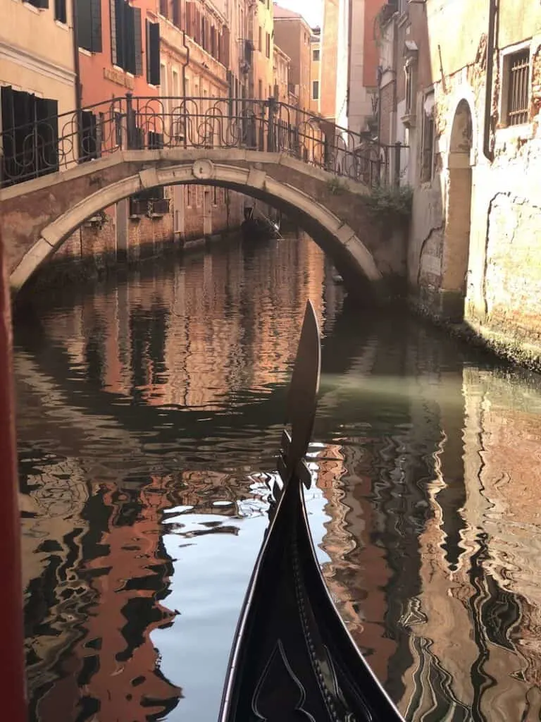 A gondola gliding on the water in Venice with a bridge to the front and just the tip of the boat visable in the image.