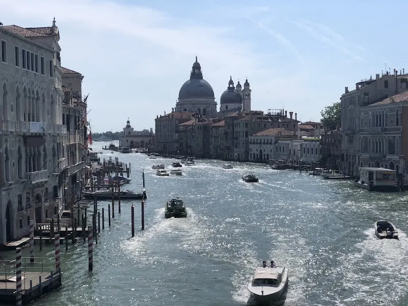 View from on top of a bridge looking towards the Grand Canal of Venice. There are several speed boats on the water below with buildings and docks to either side of the canal.