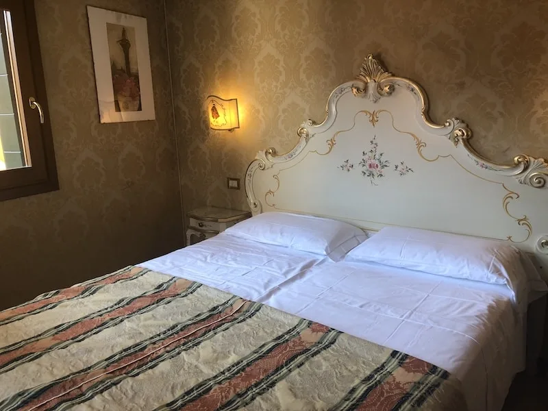 Hotel room in Venice with a queen bed that has a scroll headboard and white sheets.