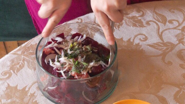 Two hands with spoons mixing beet salad in a large glass bowl.