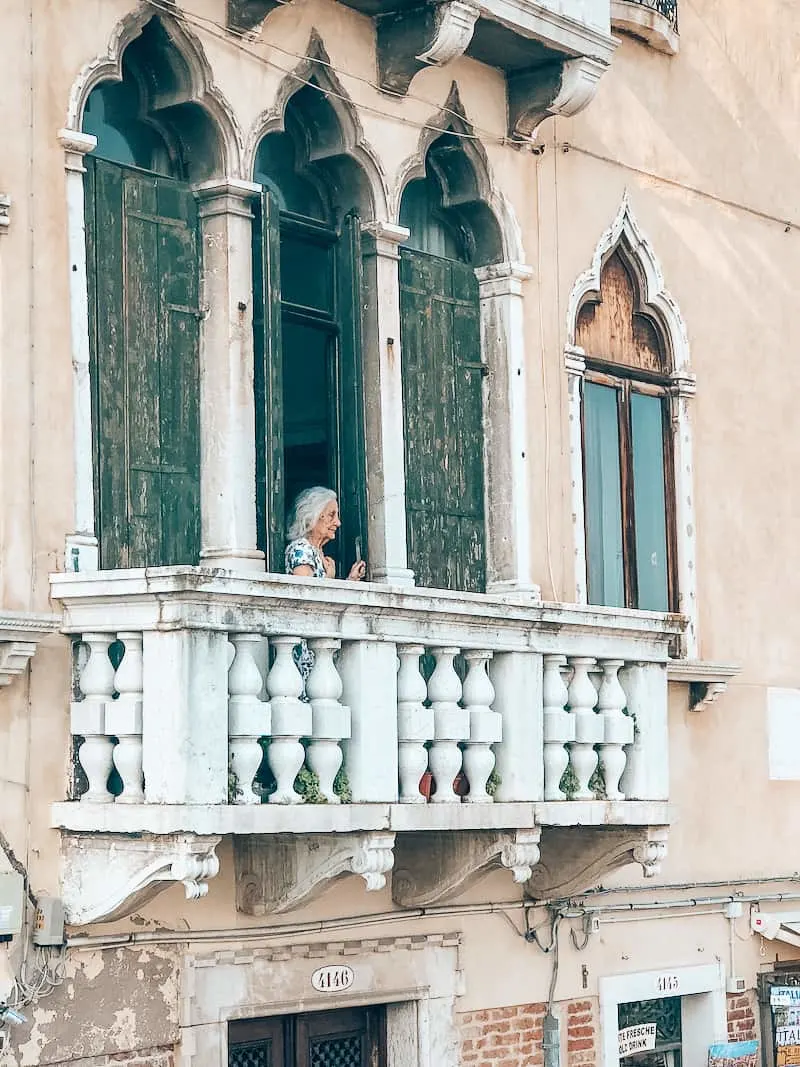 A white balcony off of an ivory building that has paint peeling. An thin, elderly woman with gray shoulder length hair looks off the balcony at the Grand Canal with her hands on the balcony railing.