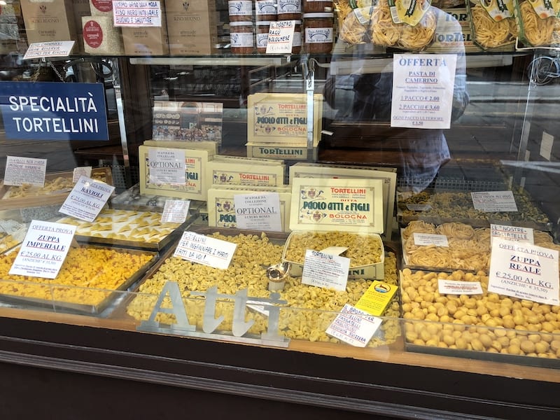 A glass window case in the front of a pasta shop. Inside the window are large trays with a variety of pastas for sale including tortellini.