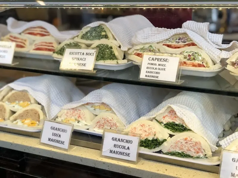 Two rows of traditional Venetian sandwiches in a bakery case. The sandwiches are soft white bread with one end open so the inside fillings are visible.