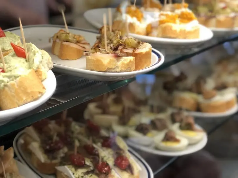 Small round pieces of bread on plates in a display case. The breads are topped with a variety of fillings such as different types of fish and vegetables.