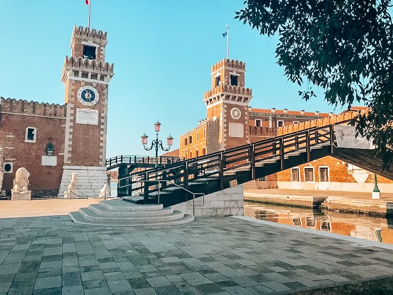 The Venice Naval Arsenal with a two tower buildings on either side of the image. A bridge crosses a canal directly in front of the buildings. A large statue of a lion is to the left of the image.