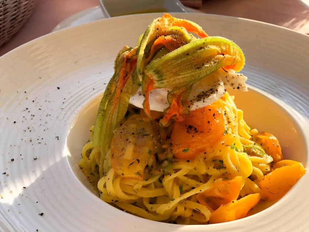 A large white bowl with a wide brim and small center area. Long spaghetti noodles are inside and topped with burrata cheese, zucchini flowers roasted tomatoes.