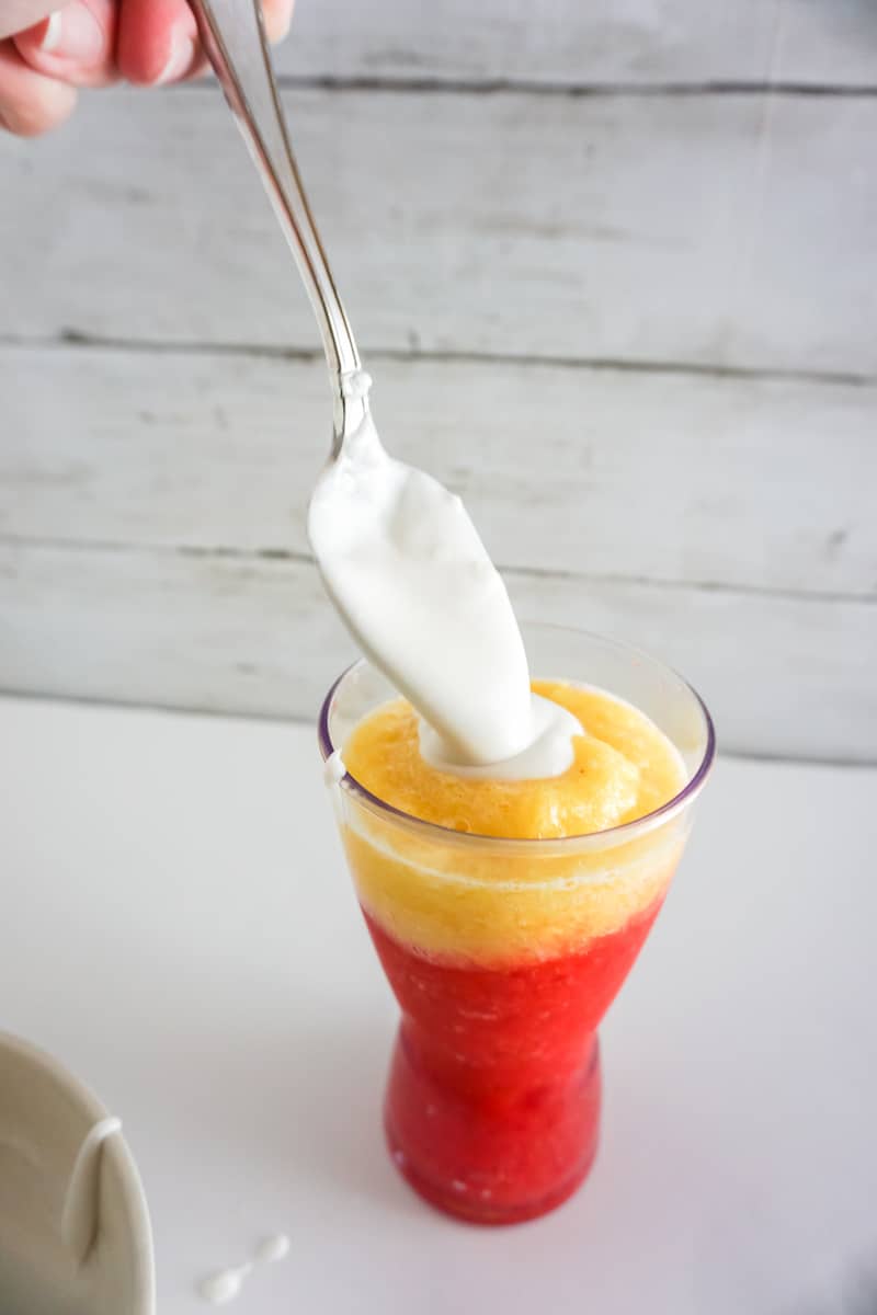 A spoon drops cream onto a glass with pineapple and strawberry puree.