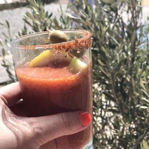 Woman's hand holding a glass with bloody mary, it has red rim salt, a lemon and olives inside