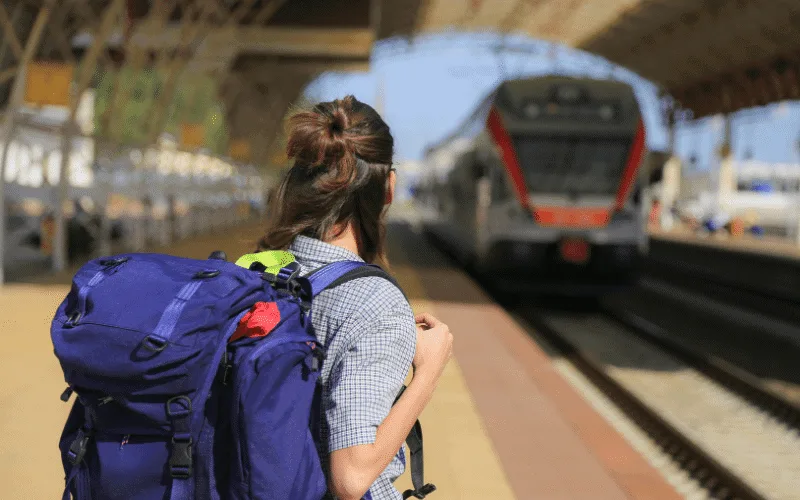 Brown haired girl waits in the foreground for a train. She has a purple backpack and is looking away from the camera.