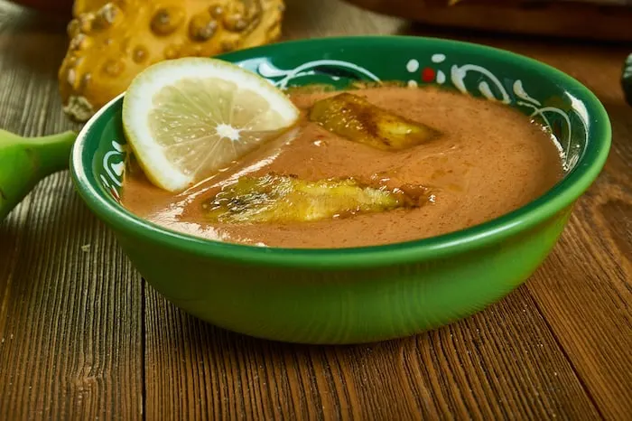 Green bowl with soup inside, plantains on the top and a lemon slice. 