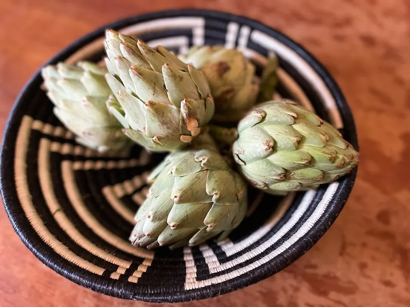 5 fresh artichokes in a black and white woven bowl on a wooden table. 