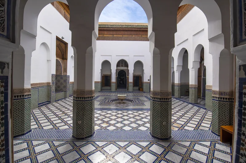 Courtyard of a Moroccan building with two white pillars in the foreground and blue and white tiled floor. 