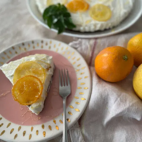 Slice of cheesecake on a pink plate with oranges and lemons to the side. It has sliced oranges on top.