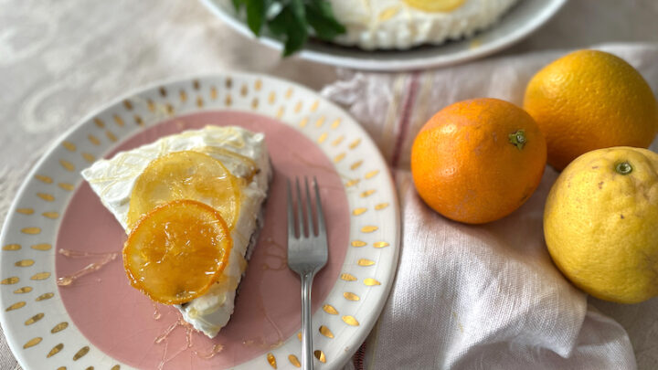Slice of cheesecake on a pink plate with oranges and lemons to the side. It has sliced oranges on top.