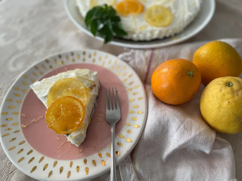 Slice of cheesecake on a pink plate with oranges and lemons to the side. It has sliced oranges on top.