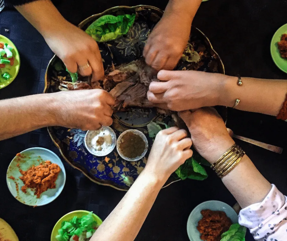 6 womens' hands reach to the middle of table that has a large plate with roasted lamb