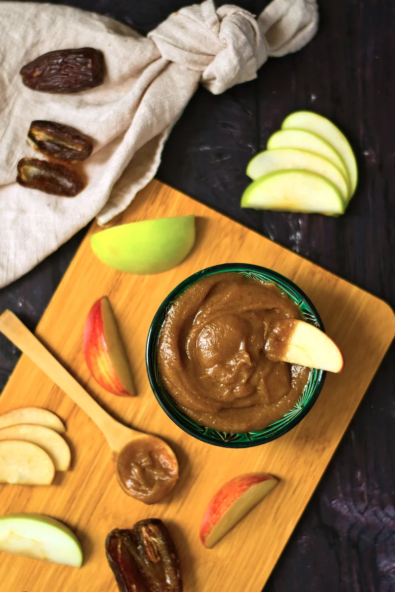 Green bowl with date caramel on a cutting board surrounded by slices of green apples