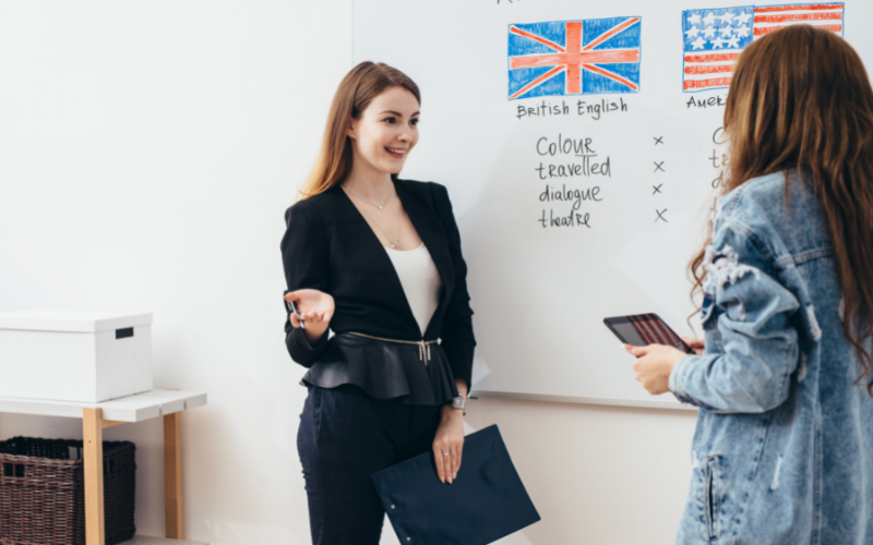 Two women stand at a white board discussing differences between American and British English