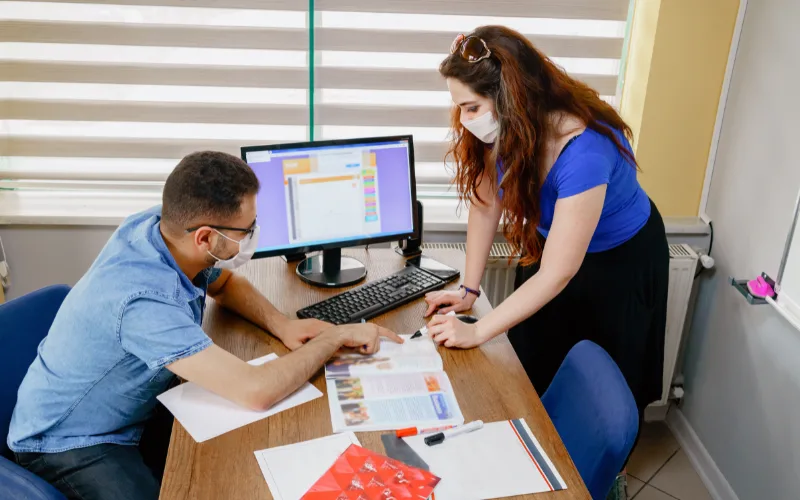 A woman stands behind a desk and a man is sitting at a desk looking at an English text.