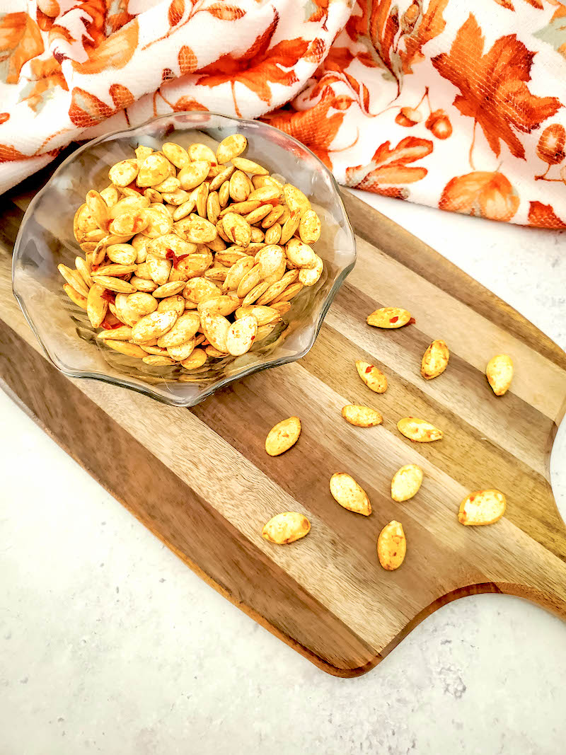 bowl of pumpkin seeds on a wooden cutting board with pumpkin seeds scattered around