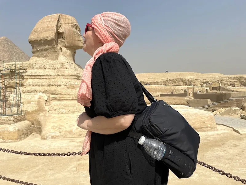 A woman in a black dress uses an optical illusion to appear as kissing the Sphinx