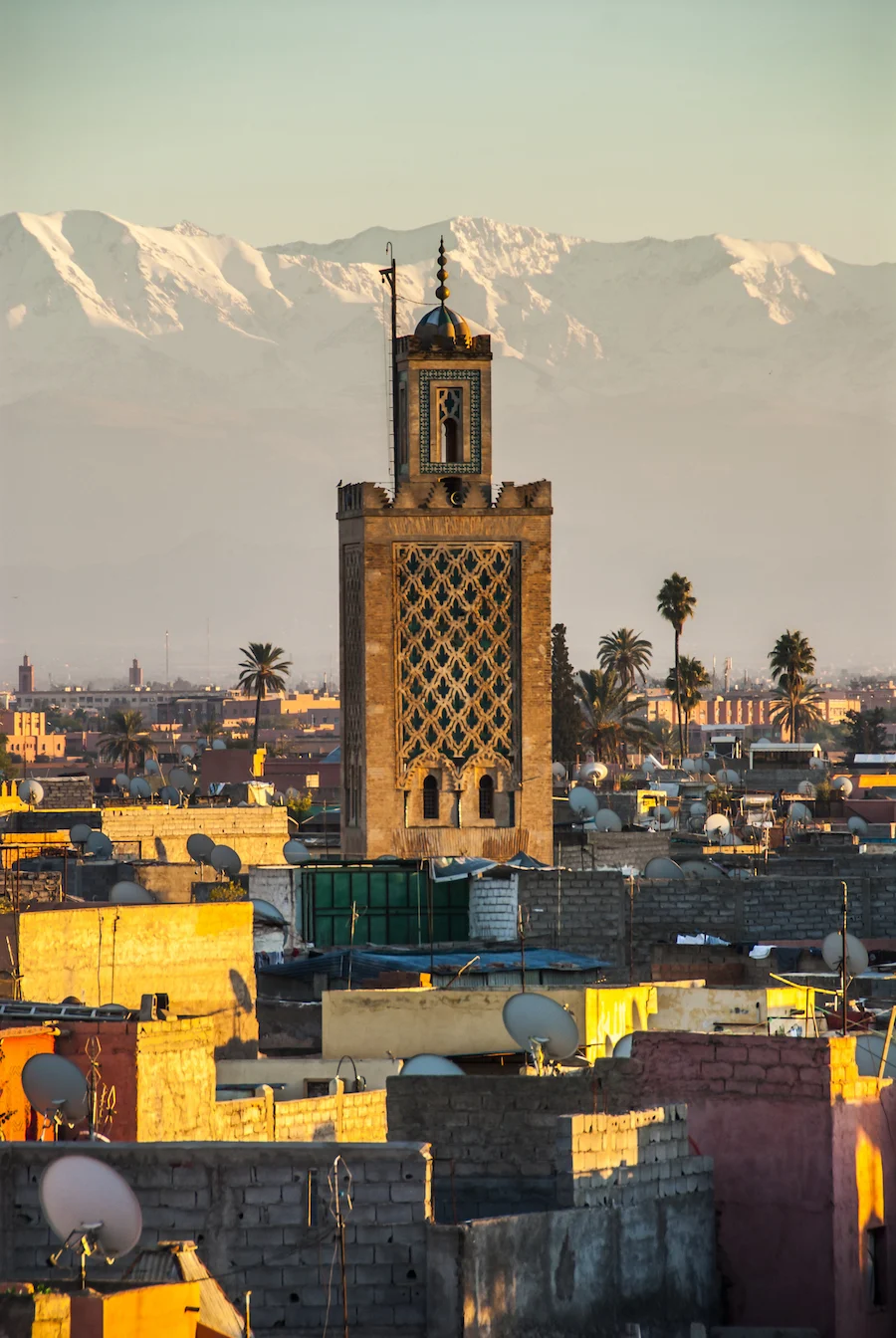 A minaret in Marrakech is in the middle with homes in front of it and snow covered Atlas Mountains in background. 